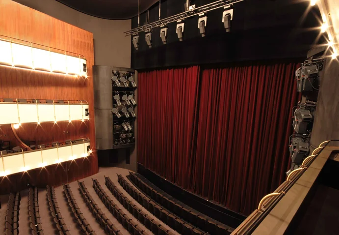 Innenraum eines Theaters mit roten Vorhängen, leeren Sitzreihen und Beleuchtung. Eine Bühne wartet darauf, sich zu öffnen.