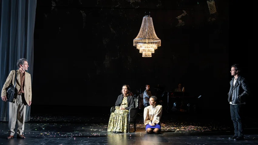 Four people on a dark theater stage with a large chandelier above. One person is seated in a chair, another on the floor.