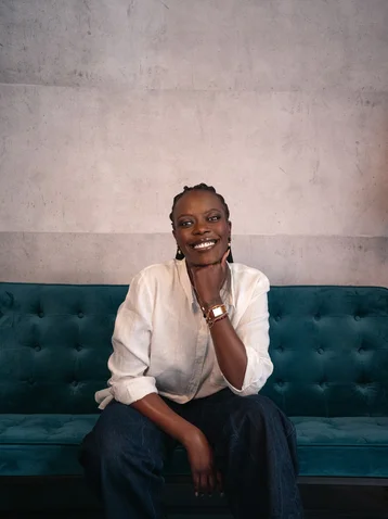 A person sits on a teal velvet sofa, smiling. They are wearing a white shirt and resting their hand on their chin.