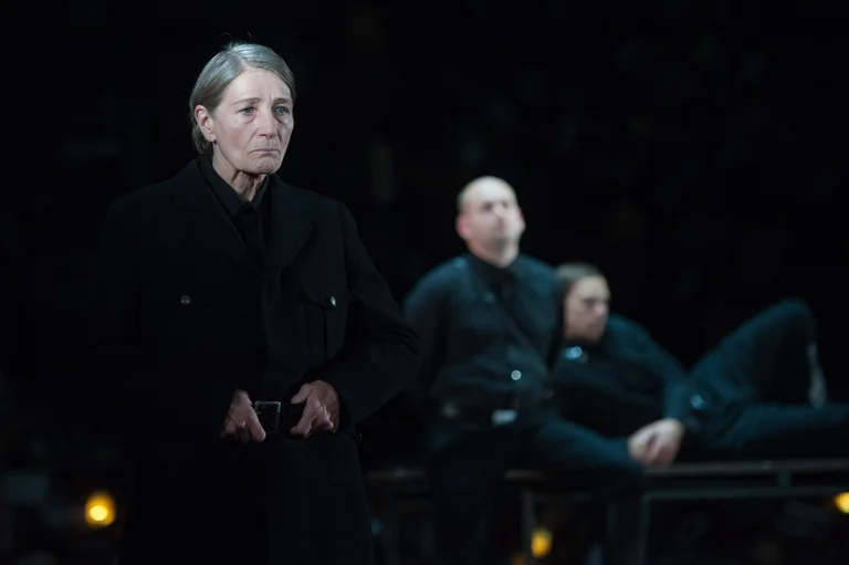 Eine Frau in schwarzer Kleidung steht im Vordergrund eines Theaterstücks, während zwei Personen im Hintergrund sitzen und liegen.