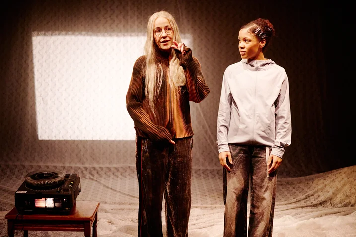 Two women stand on stage. An older woman speaks into a microphone while the younger woman listens attentively.