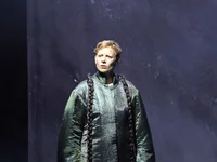 A person stands on a dimly lit stage wearing a voluminous, long-sleeved costume with long braids, creating a dramatic effect.