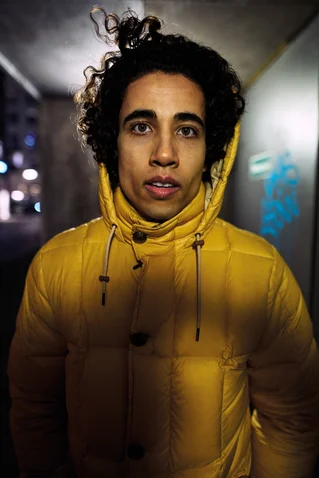 Ein Mensch mit lockigem Haar trägt eine gelbe Daunenjacke. Er steht draußen, vermutlich in einer städtischen Umgebung bei Nacht.