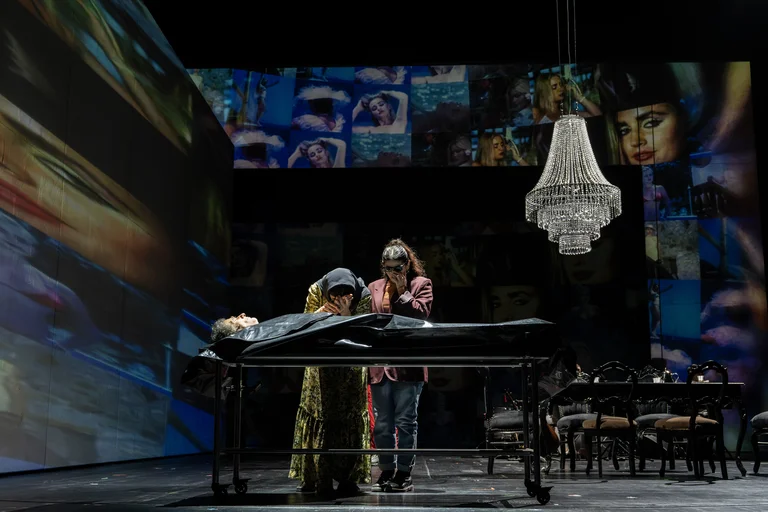 Szene aus einem Theaterstück mit einer liegenden Person auf einem Tisch, umgeben von zwei weiteren Personen in einem dunklen Raum.