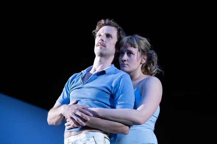 A man and a woman in blue clothing stand on stage. The woman embraces the man from behind.