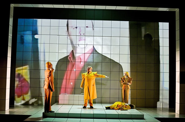 Three actors in a dramatic stage scene with a large projected image in the background. One figure gestures dramatically.