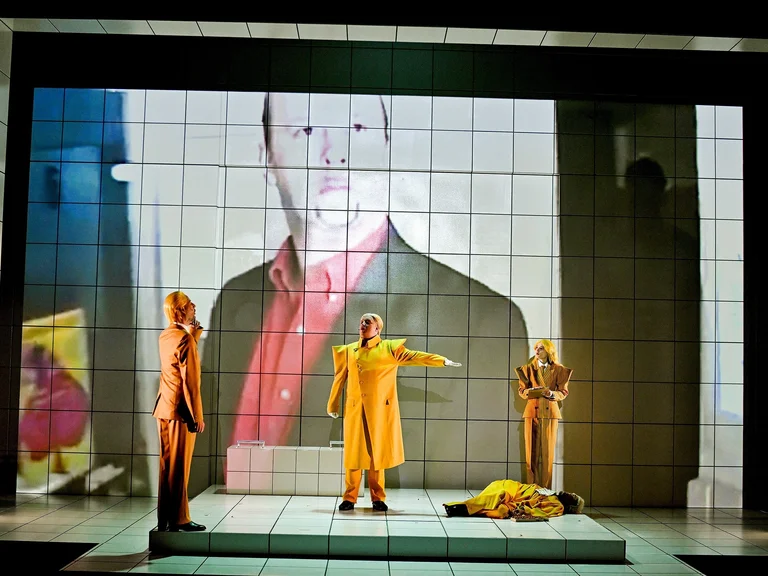 Drei Schauspieler in einem dramatischen Bühnenbild mit einem großen projizierten Bild im Hintergrund. Eine Figur zeigt dramatisch.