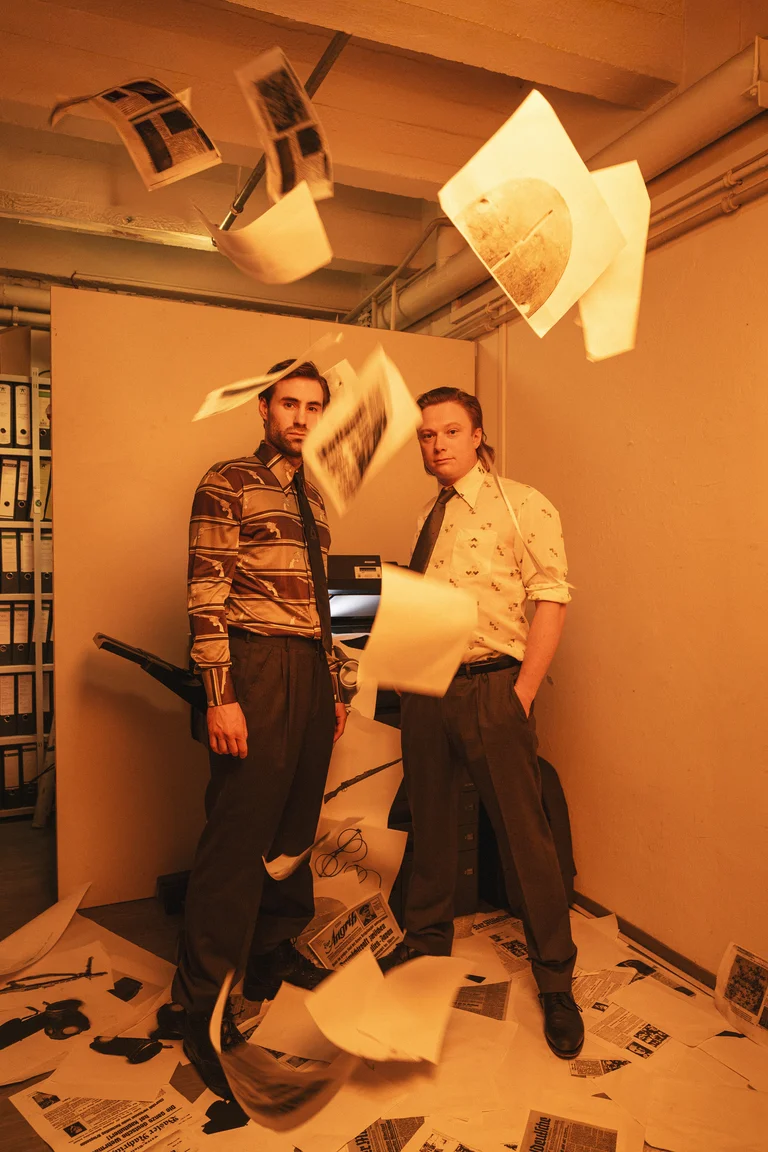 Two men stand in an office with papers falling around them. A printer and a shelf are visible in the background.