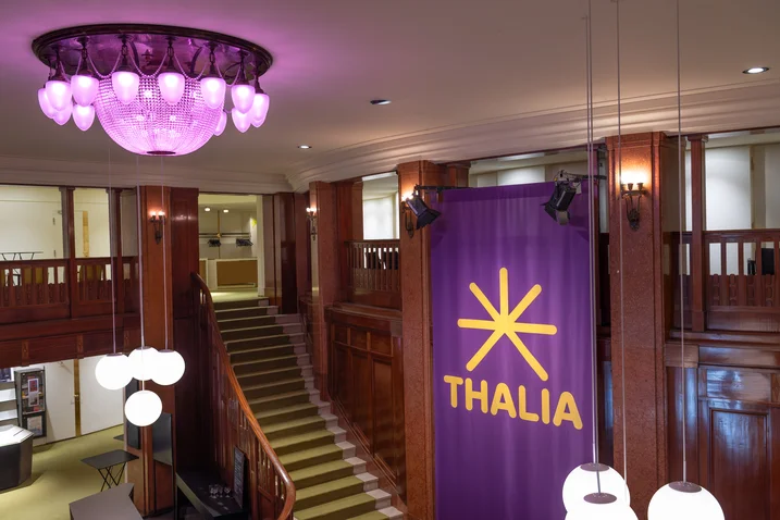 A stylish foyer featuring a large purple chandelier. A banner with "THALIA" is hanging next to a staircase.