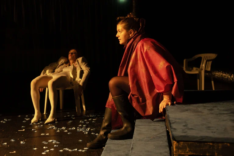 Eine Schauspielerin in einem roten Mantel sitzt auf einer Bühne. Zwei Personen in weißen Anzügen entspannen sich auf Stühlen.