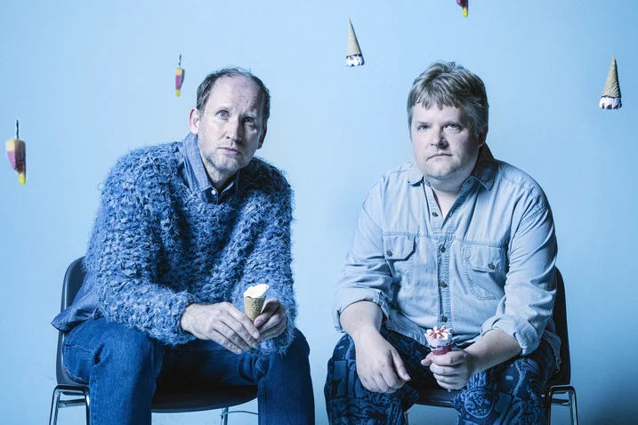 Two men sit on chairs against a blue background. They hold ice cream cones and wear casual clothing.