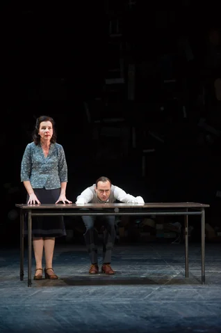 Two people on stage, a woman and a man, with a table between them. They appear focused and engaged.