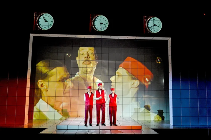 Three people in red outfits stand on stage. Three large clocks and faces are projected in the background.