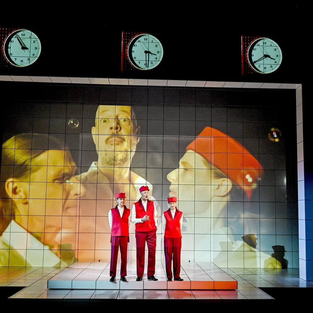 Drei Personen in roten Outfits stehen auf einer Bühne. Im Hintergrund sind drei große Uhren und Gesichter projiziert.