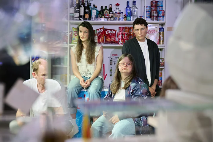 A group of four people sits casually in a grocery store in front of shelves with snacks and drinks.