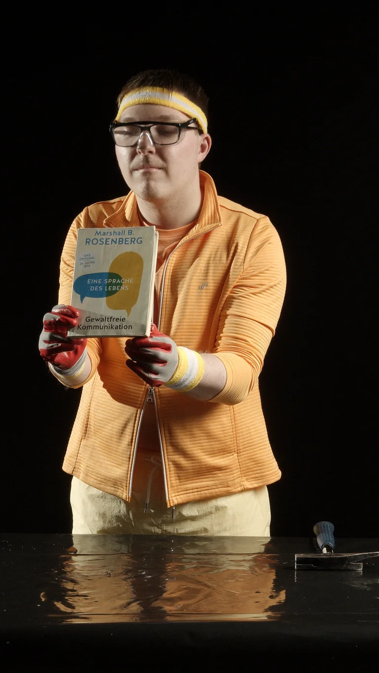 Eine Person in orangener Kleidung hält ein Buch hoch. Der Hintergrund ist dunkel. Die Szene wirkt künstlerisch inszeniert.