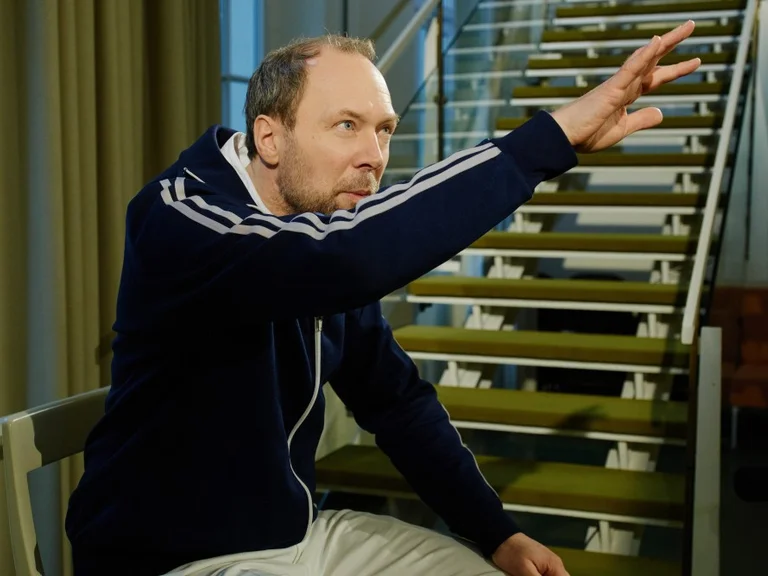 Ein Mann in Freizeitkleidung sitzt auf einer Treppe und hebt die Hand, als ob er etwas erklärt.