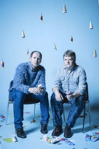 Two men sit in a blue room on chairs, holding ice cream cones, with cones hanging from the ceiling and paper scattered.