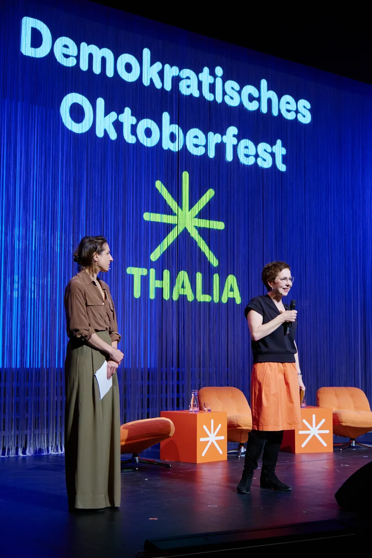 Sonja Anders und Laura de Weck stehen auf einer Bühne beim "Demokratisches Oktoberfest" im Thalia. Hintergrund zeigt Schriftzug und orangene Dekoration.