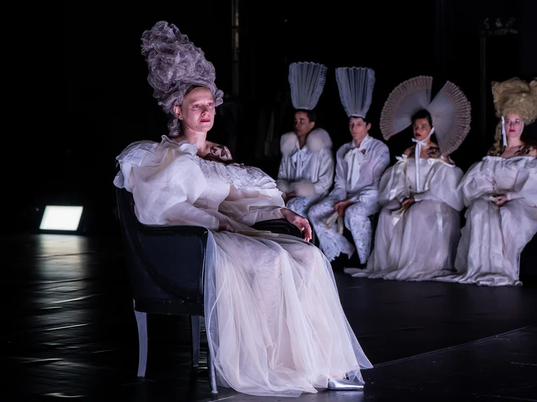 Eine Gruppe von Darstellern in aufwändigen, weißen Kostümen und hohen Frisuren sitzt auf einer Bühne, in einem Theater.