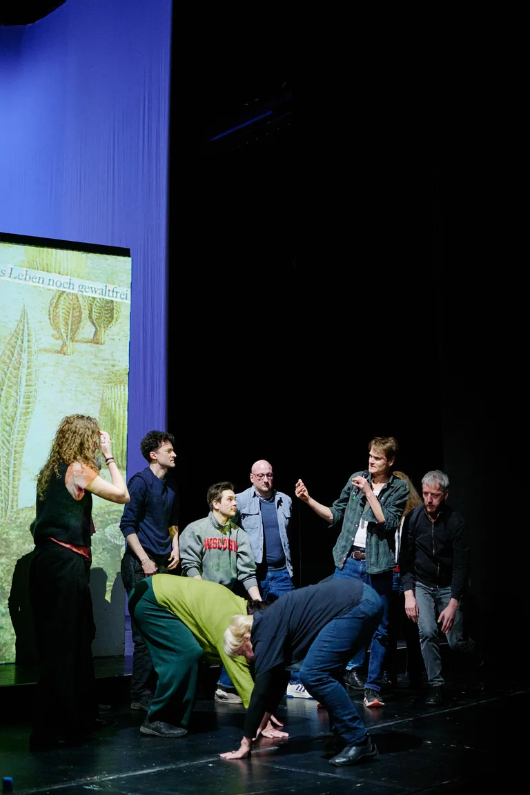 Eine Gruppe von Menschen auf einer Bühne, einige in dynamischen Posen. Hintergrund ist eine Leinwand mit Illustrationen.