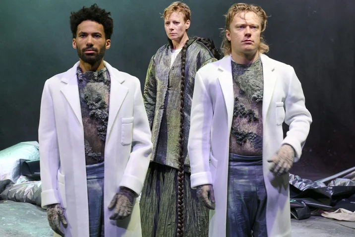 Drei Menschen in futuristischen Outfits stehen auf einer Bühne, die mit dramatischem Licht und dunklem Hintergrund beleuchtet wird.