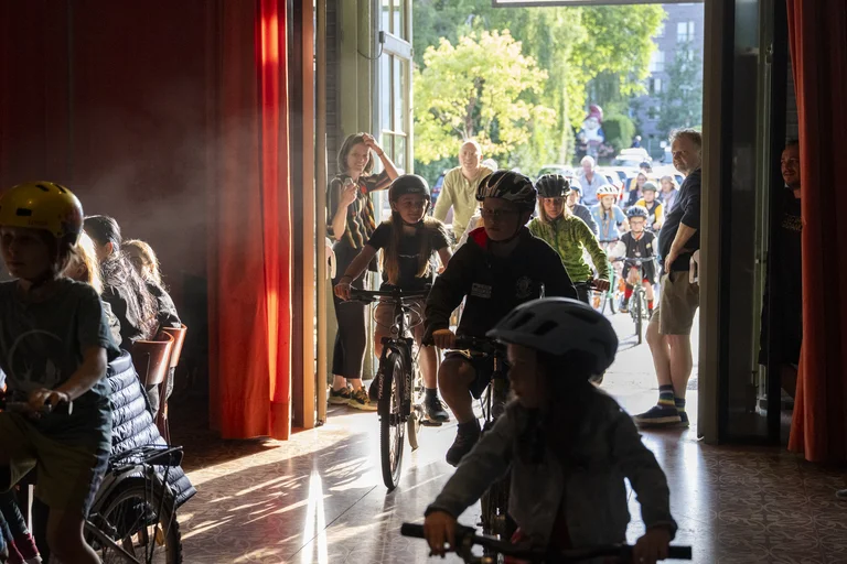 Kinder fahren auf Fahrrädern in einen Raum mit roten Vorhängen hinein. Sonnenlicht scheint durch offene Türen.
