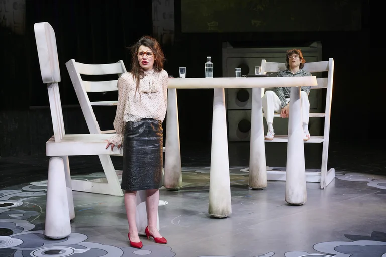 Eine Frau steht in einem grauen Rock und roten Schuhen vor riesigen Möbeln auf einer Bühne. Eine andere Person sitzt am Tisch.