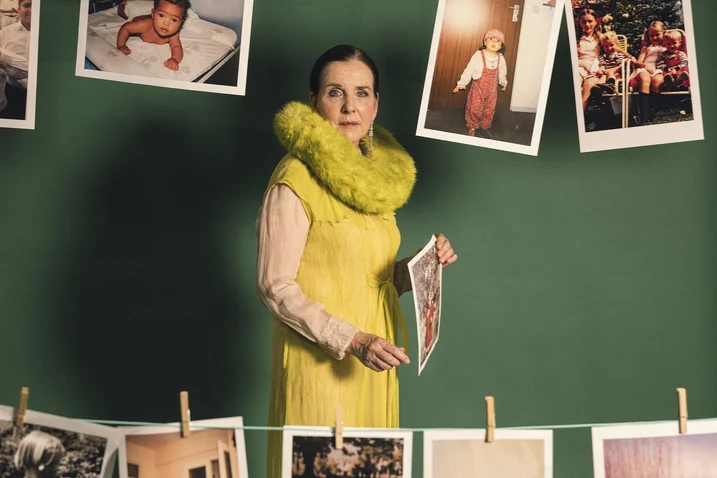 A woman in a yellow dress stands in front of a green wall with hanging photos. She holds a picture.
