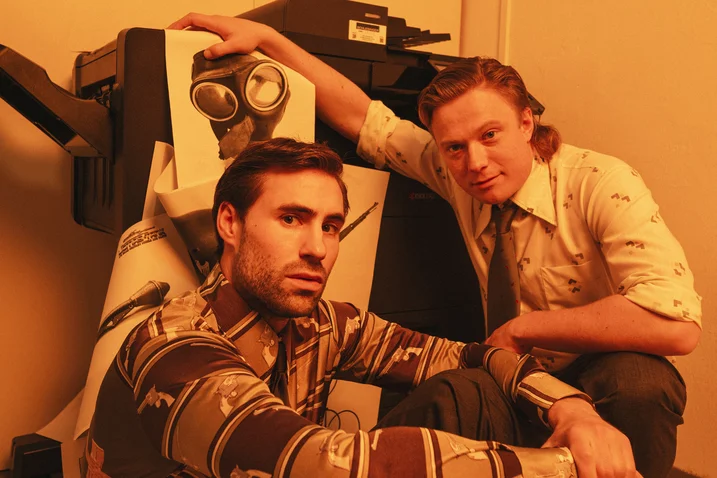 Two men in retro outfits pose beside a quirky object with glasses, in a room bathed in orange light.