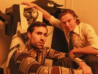 Two men in retro outfits pose beside a quirky object with glasses, in a room bathed in orange light.