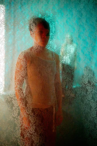 Ein unscharfes Bild einer Person hinter transparentem Stoff mit floralen Mustern. Die Farben sind Blau und Orange.