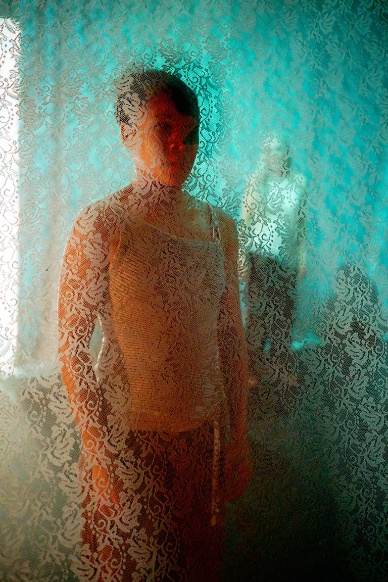 Ein unscharfes Bild einer Person hinter transparentem Stoff mit floralen Mustern. Die Farben sind Blau und Orange.