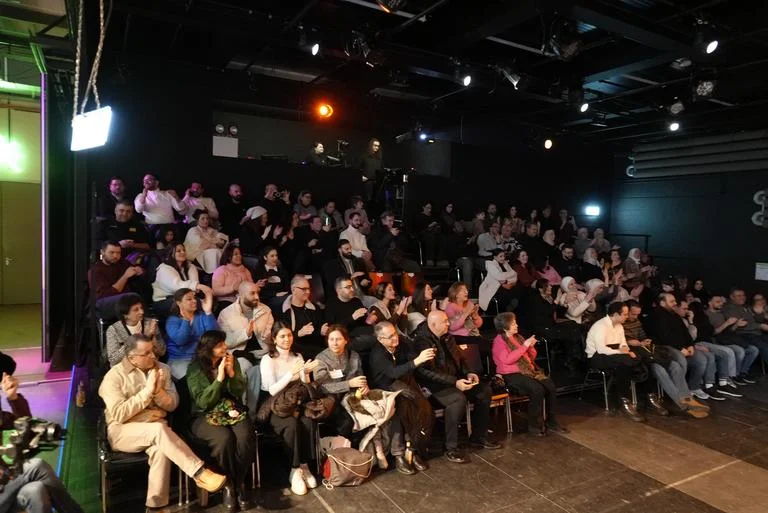 Menschenmenge sitzt in einem dunklen Theaterraum, aufmerksam auf die Bühne schauend. Die Atmosphäre ist gespannt und interessiert.