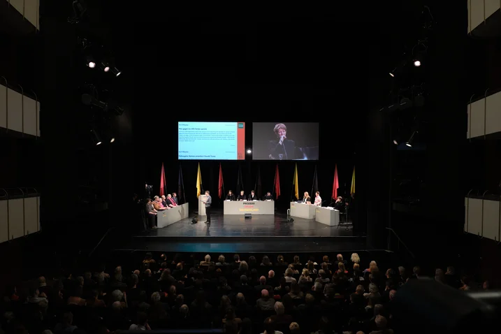 Ein Auditorium mit einer Bühne, auf der Leute an Podiumstischen sitzen. Im Hintergrund sind Bildschirme und Fahnen zu sehen.