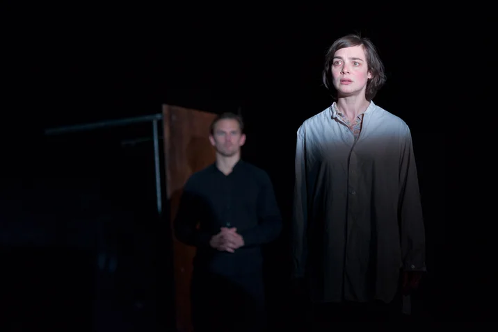 Two individuals stand in a dark room, one illuminated in the foreground. Both look attentively into the distance.