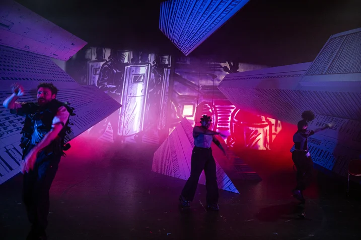 Dancers perform in front of a futuristic set with colorful lights and geometric shapes. The atmosphere is dynamic and energetic.
