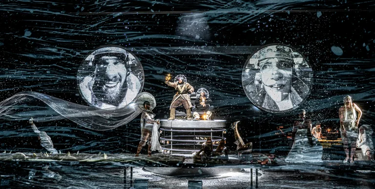 Eine düstere Theaterinszenierung mit komplexem Bühnenbild, umgeben von zwei großen, schwarz-weißen Porträts. Schauspieler in dramatischen Posen.