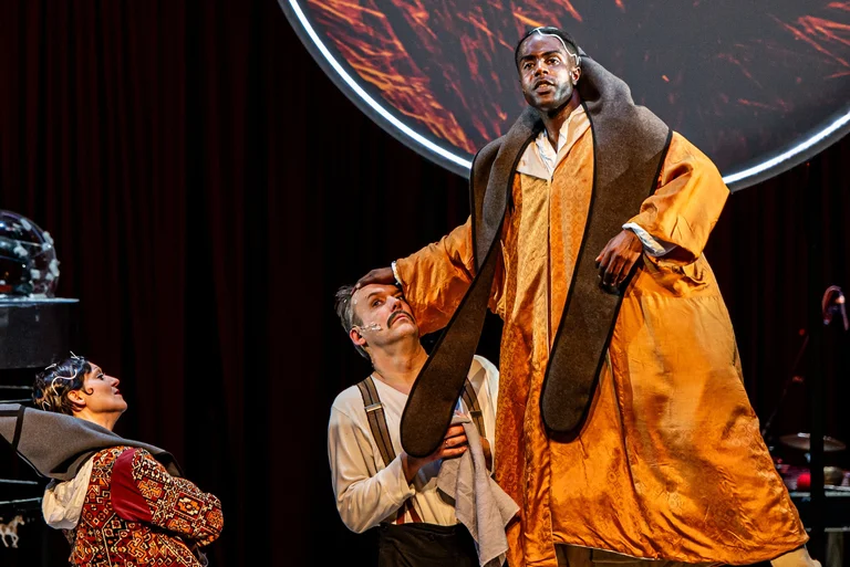 Drei Schauspieler in historischen Kostümen auf der Bühne. Einer trägt ein prächtiges, goldenes Gewand und wird dramatisch beleuchtet.