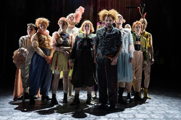 Eine Gruppe von Menschen in fantasievollen Kostümen steht auf einer Bühne. Sie tragen bunte, dynamische Kleidung und scheinen bereit für eine Aufführung.