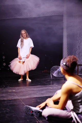 Zwei Tänzerinnen in einem Probenraum. Eine trägt ein Tutu und T-Shirt, während die andere sitzt. Lichteffekte im Hintergrund.