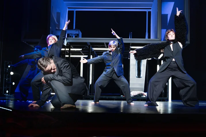 Four dancers in dark suits energetically perform on stage. Their poses are dynamic and expressive, resembling a modern theater piece.
