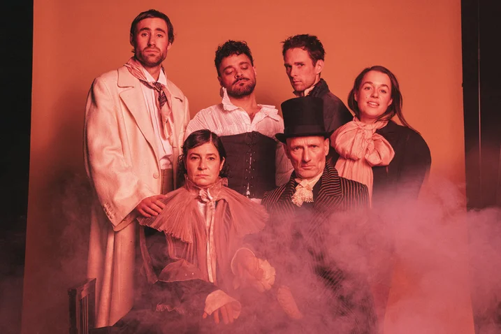 A group of six people poses in historical costumes with fog effects, set against an orange background. They appear serious.