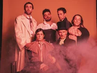A group of six people poses in historical costumes with fog effects, set against an orange background. They appear serious.