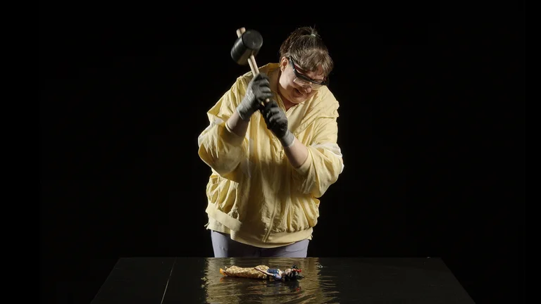 Eine Person in gelbem Pullover schlägt mit einem Hammer auf eine Oberfläche. Trägt Schutzbrille und Handschuhe. Schwarzer Hintergrund.