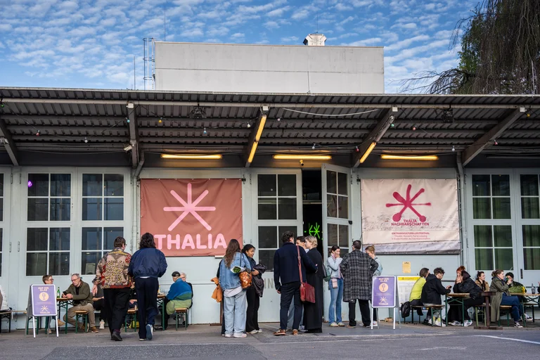 People gather outside a building with Thalia banners. Some are standing and chatting, while others are seated on chairs.