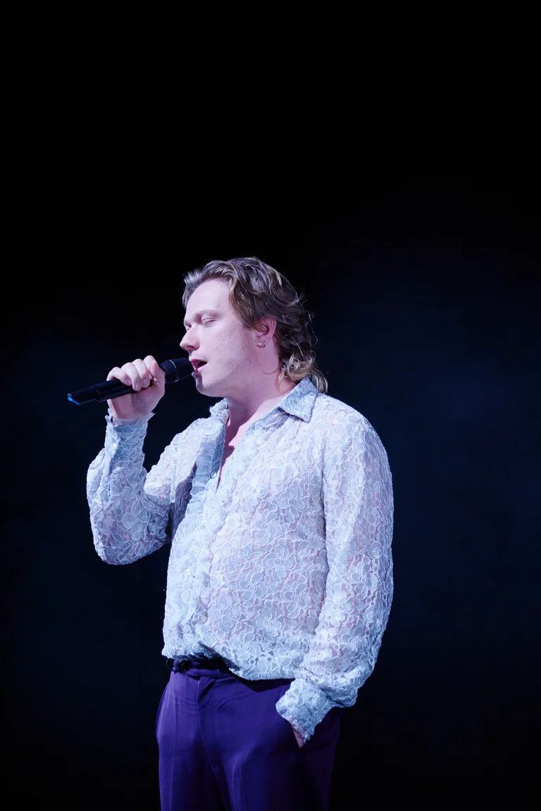 Jannik Hinsch in einem weißen Spitzenhemd hält ein Mikrofon und singt auf einer Bühne vor dunklem Hintergrund.