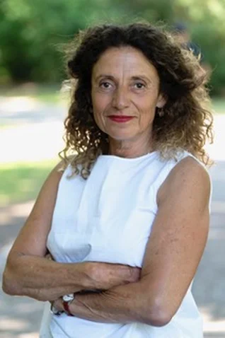 Adriana Altaras with curly hair wears a sleeveless white top, standing outdoors with arms crossed. Background is blurred greenery. (portrait picture)