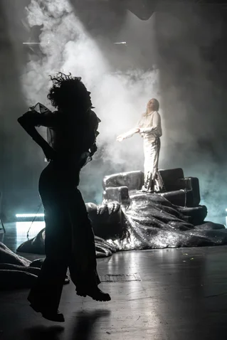 A dramatic stage scene with a person in light in the background, while a silhouetted figure dances in the foreground.