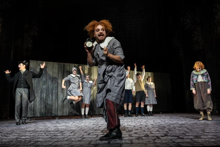 A theatrical performance with various characters on stage. A person in a bold costume is in the foreground.